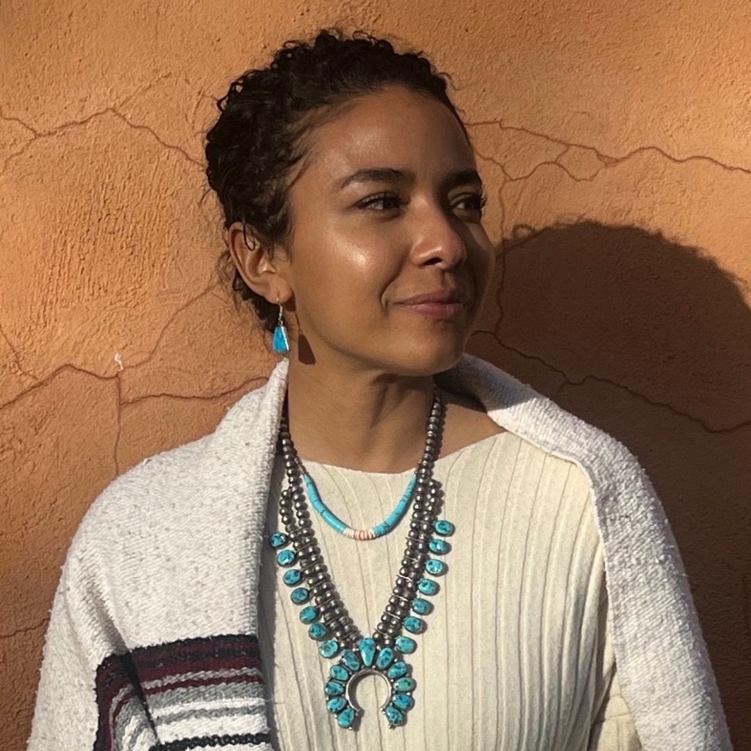 Deoné Newell in front of an adobe wall in warm sunlight, wearing turquoise jewelry and a woven shawl
